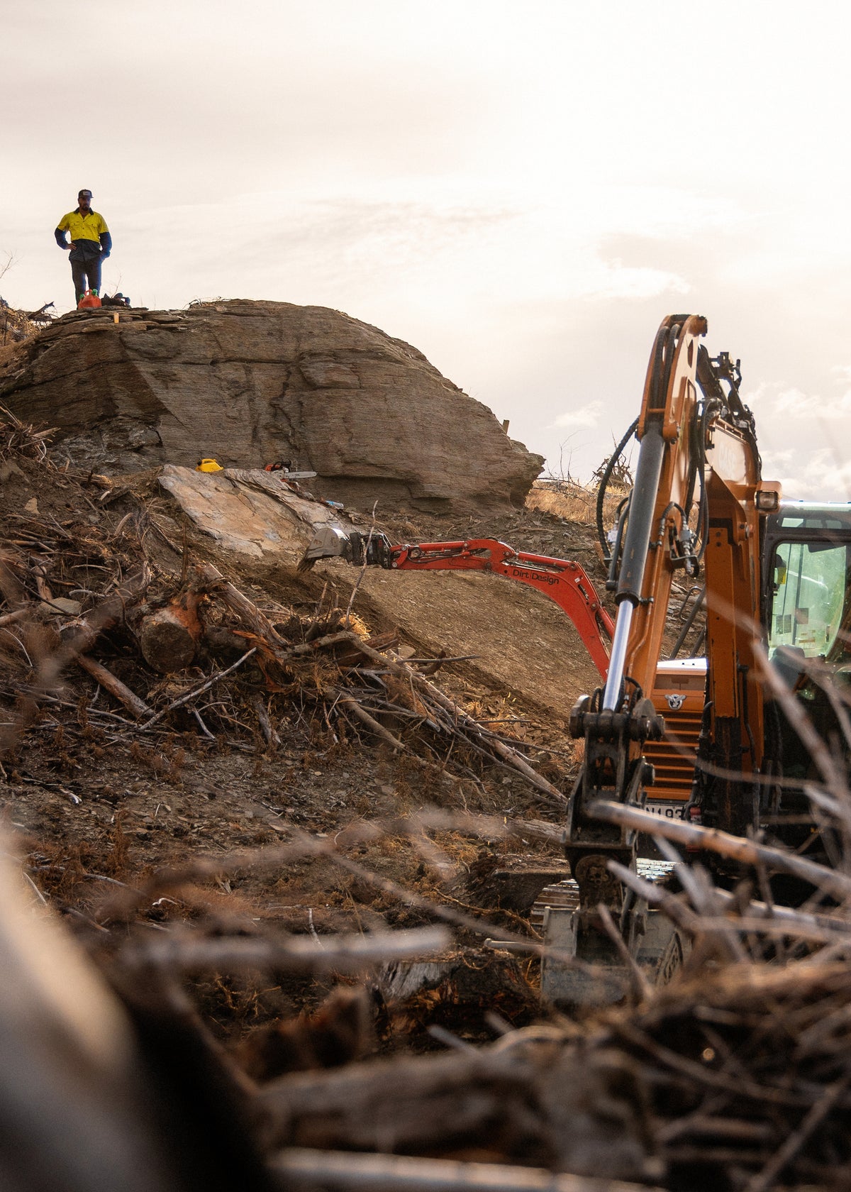 An orange excavator is shown in a construction or demolition site, with a person standing on a large rock formation in the background under an overcast sky.