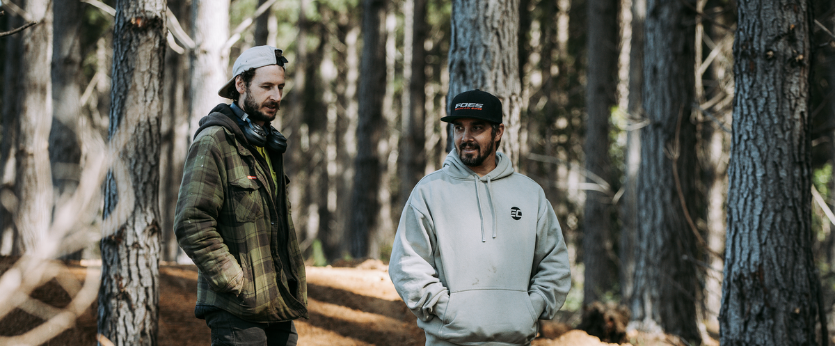 Two men standing on a forest path surrounded by tall trees, one wearing a green plaid jacket and a white cap, and the other wearing a light gray hoodie and a black cap. Sam hill signature trail builders, Sam and Dave